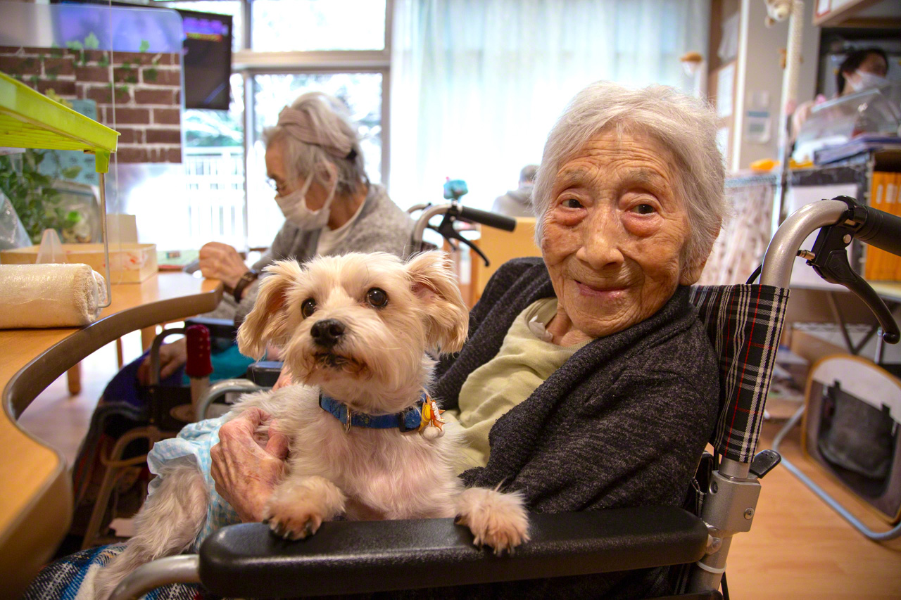 Ms. Nozawa, a 97-year-old resident, enjoys a quiet moment with her dog Mick as care staff look after the day’s tasks. (© Ōnishi Naruaki)
