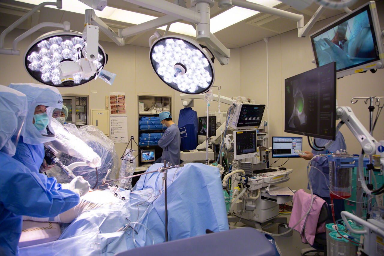 The Mako robotic-arm assisted surgery system springs into action. Dr. Matsubara guides the robotic-arm as the attachment spins, removing bone tissue. The monitor at right shows the section to be removed in green. A clean removal leaves only white. Any excess removal appears in red. If the cutting proceeds more than 2 mm beyond the predetermined boundary, the arm automatically stops. (© Ōnishi Naruaki)