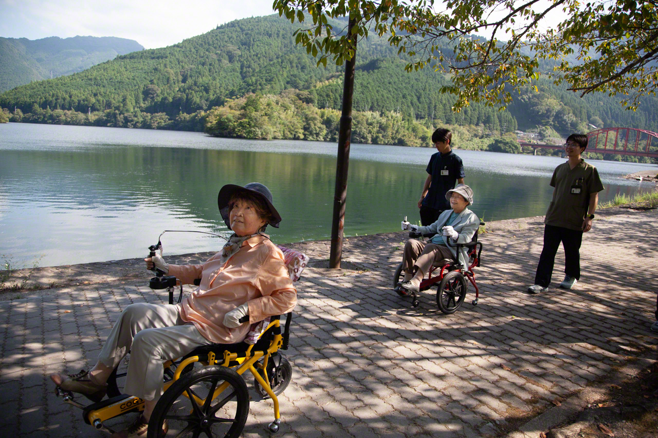 Two friends from Daycare Hikari 2, aged 94 and 83, take the air around Lake Kinsha in their Cogy wheelchairs, which they have been using for about six years. “I used to have to go to the doctor twice a week to get shots for knee pain,” says one. “Now I don’t go at all. The Cogy has made me so much better.” (© Ōnishi Naruaki)