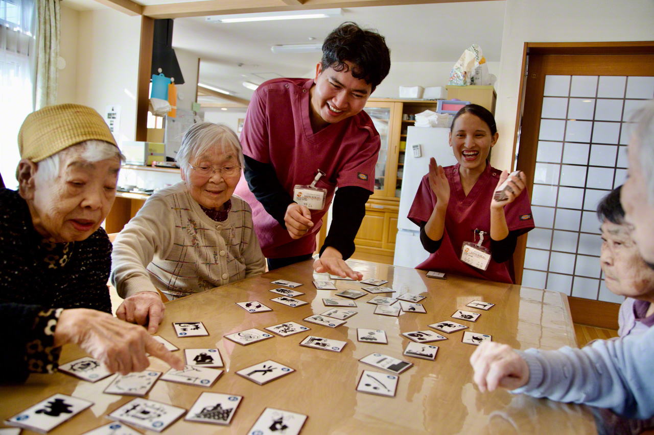 Taman applaudit tandis que Sam, debout, aide les joueurs à choisir la bonne carte dans une partie de karuta.